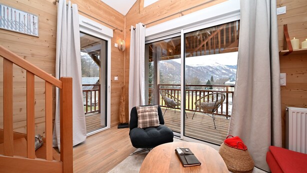 Een houten interieur met een schuifdeur naar een balkon, een zwarte stoel met een geruit kussen en een houten tafel met een afstandsbediening.