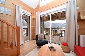 A wooden interior with a sliding glass door leading to a balcony, a black chair with a plaid pillow, and a wooden table with a remote control. - Ideal Getaway at Lake Loudenvielle – Comfortable Apartment with Outdoor Activities (Genos)