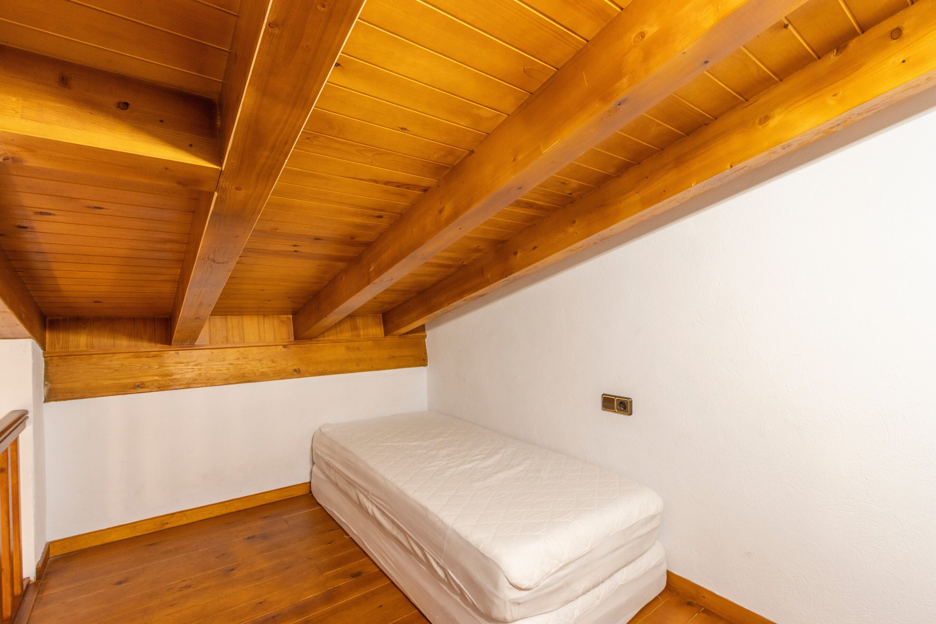 Una stanza piccola con un letto singolo, travi in legno a vista al soffitto, pareti bianche e pavimento in legno.