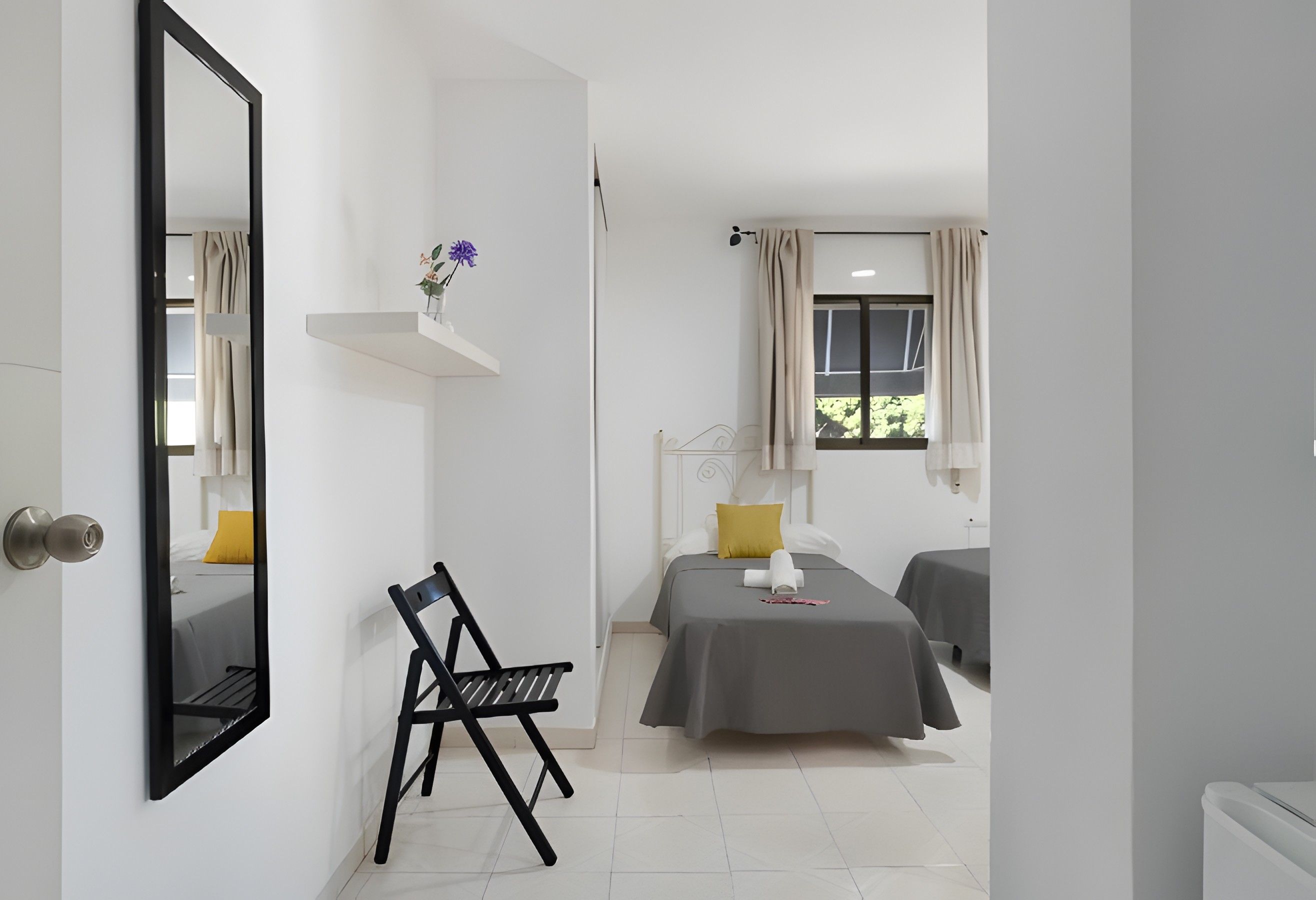 Una habitación de hotel con una cama, una silla, una mesita de noche, un espejo y una ventana con cortinas.