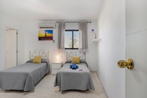 A room with two beds, a window with curtains, an air conditioner, a nightstand with a lamp, and a wall-mounted shelf with a vase.