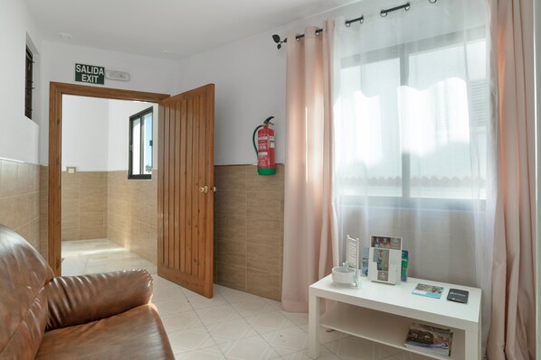 A room with a wooden door marked 'SALIDA EXIT,' a white shelf with magazines, a fire extinguisher, and a window with sheer curtains.