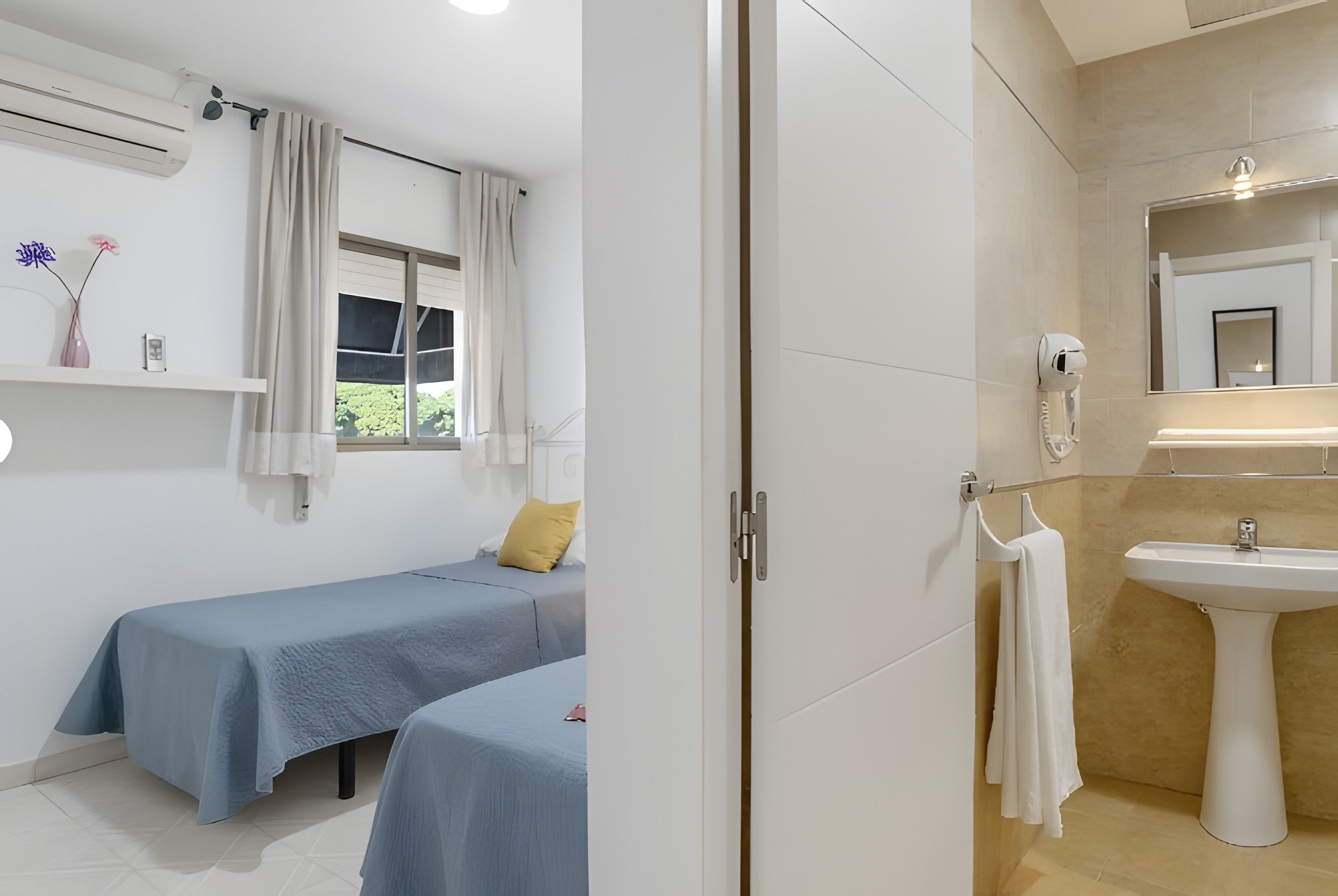 Une chambre avec deux lits, une fenêtre avec des rideaux, un climatiseur fixé au mur et une salle de bain avec un lavabo et un miroir.