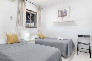 A hotel room with two beds, a nightstand, a chair, a window with curtains, a picture on the wall, and a shelf above the bed.