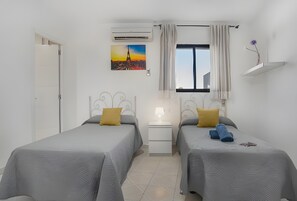 Een hotelkamer met twee bedden, een raam met gordijnen, een schilderij van een stadsgezicht en een airconditioner.