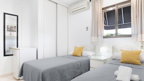 A hotel room with two beds, a window with curtains, an air conditioner, a bedside table with a lamp, and a mirror.