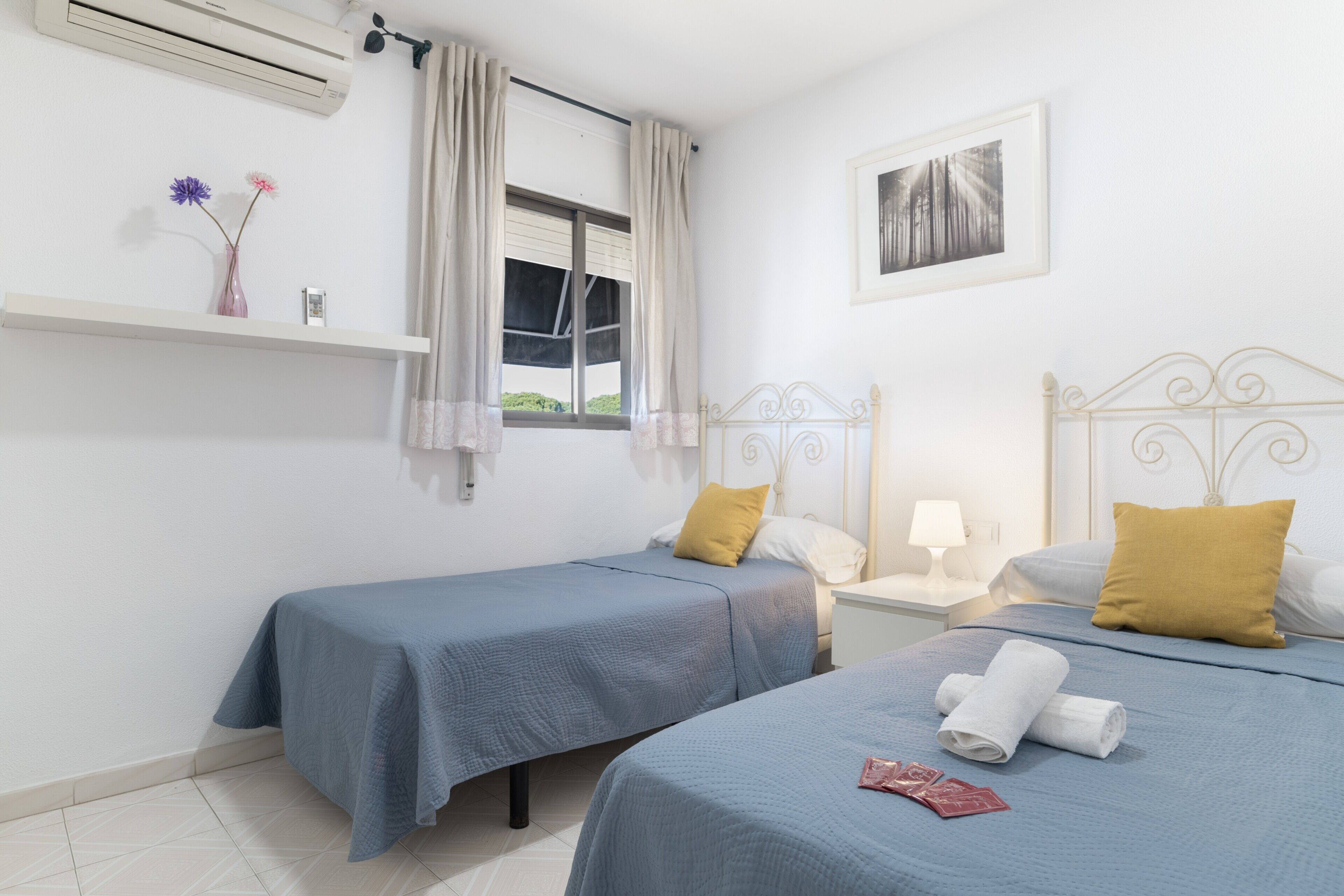 Camera d'albergo con due letti singoli, un comodino con una lampada, una finestra con tende, una mensola con fiori e un quadro appeso al muro.