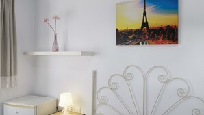 Une chambre à coucher avec un lit, une table de chevet, une lampe, un vase avec des fleurs, une image de la Tour Eiffel et un petit réfrigérateur.