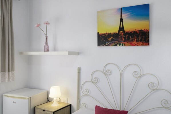 A bedroom with a bed, a nightstand, a lamp, a vase with flowers, a picture of the Eiffel Tower, and a small refrigerator.