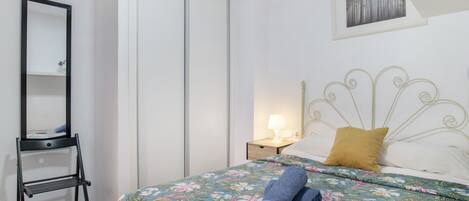 Een slaapkamer met een bed, nachtkastje, lamp, spiegel en een garderobe.