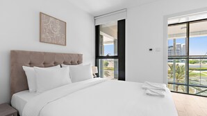 A modern hotel room with a large bed, a headboard, a framed artwork, a nightstand, and a view of the city through large windows. - Silkhaus Al Raha Lofts 2 (Abu Dhabi)