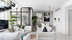A modern living room with a grey sofa, a round coffee table, and a TV. There are plants, a painting, and a staircase visible. - Silkhaus Al Raha Lofts 2 (Abu Dhabi)