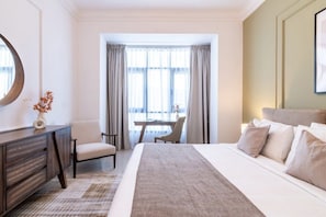 A hotel room with a bed, a desk, a chair, and a view of the outdoors through large windows.