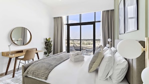 Ein Hotelzimmer mit einem großen Bett, einem Schreibtisch, einem Sessel, einem runden Spiegel und Blick auf die Stadt durch große Fenster.