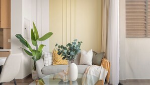 Una sala de estar moderna con una mesa de centro de vidrio, un sofá con cojines, una planta en maceta y una ventana con persianas.