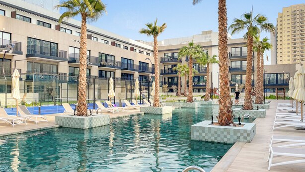 Un área de piscina con sillones de descanso y palmeras, rodeada de edificios modernos.