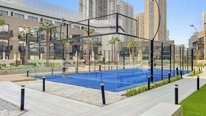 Una cancha de tenis con red, rodeada por una cerca, y contigua a una zona de piscina con sillas de descanso y sombrillas.