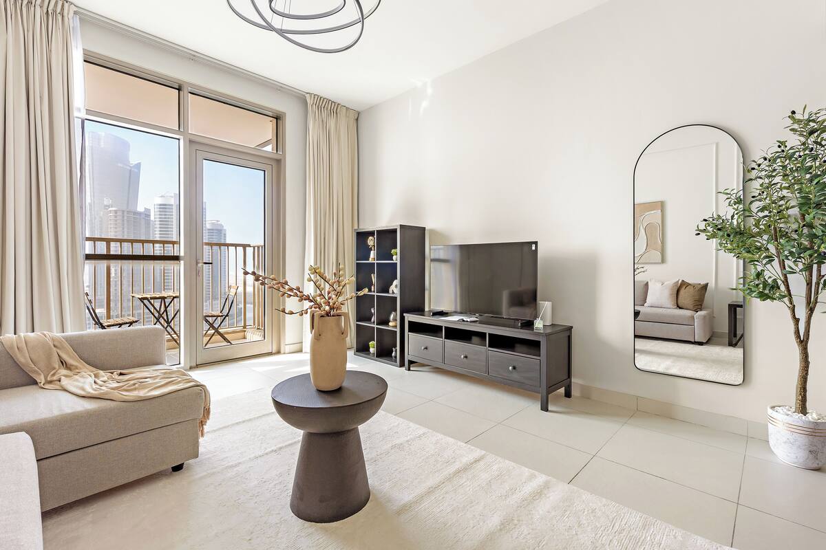 A modern living room with a sofa, a round table with a vase, a TV stand, and a large mirror.