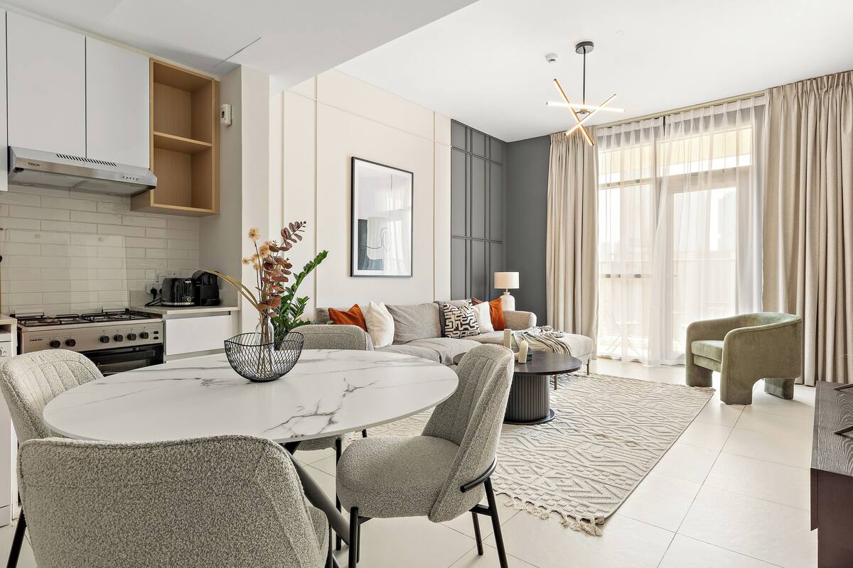 A modern kitchen with a gas stove, a built-in oven, and a stainless steel range hood. There is a dining area with a marble table and upholstered chairs. The living room features a sofa set, a coffee table, and a large window with curtains.