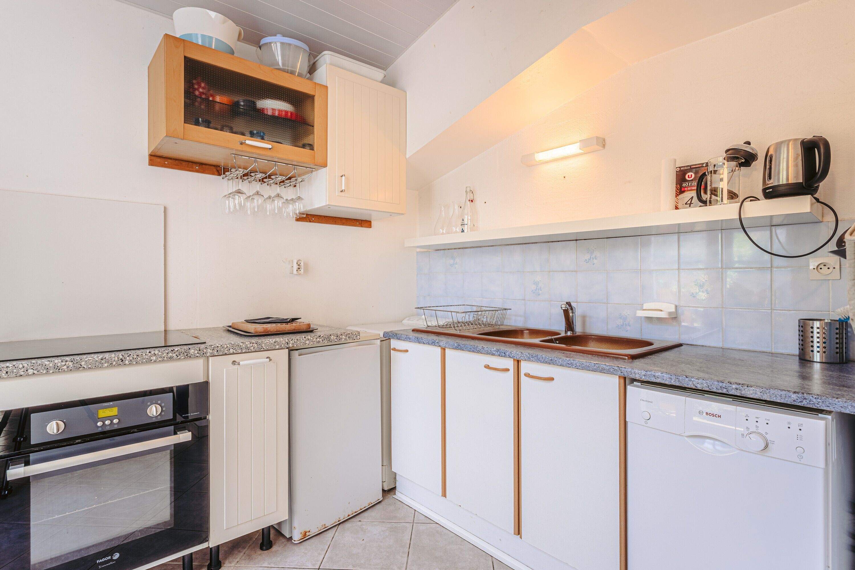 Una cocina con horno empotrado, lavavajillas y un armario de madera con copas de vino colgadas.