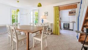 Un comedor con mesa de madera y sillas blancas, una lámpara azul y una cocina al fondo.