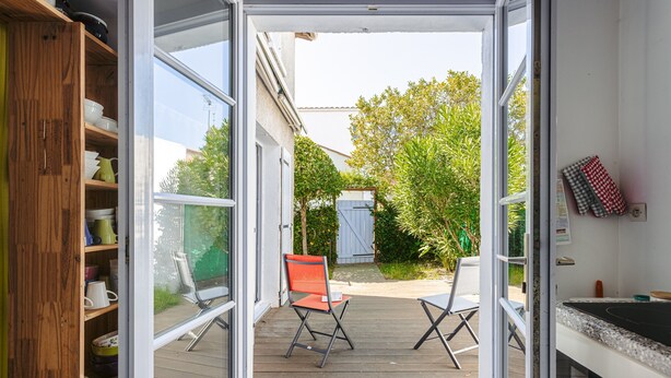 Puerta corredera abierta que da a una terraza de madera con dos sillas, una mesa y una zona de cocina con mostrador.