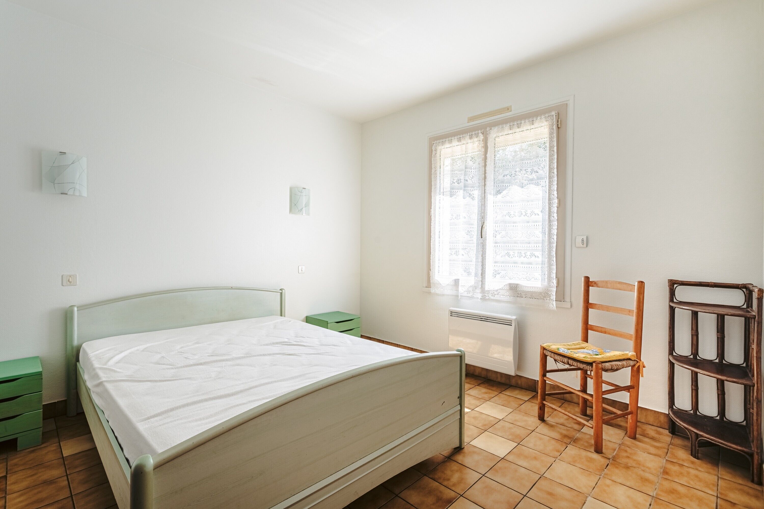 A bedroom with a bed, a chair, a nightstand, a radiator, and a window with a curtain.