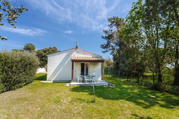 A small, single-story house with a tiled roof, a patio with outdoor furniture, and a well-maintained lawn. - Charming Detached House with Terrace, Garden, and Garage 5 Minutes from the Beaches (Saint-Trojan-les-Bains)