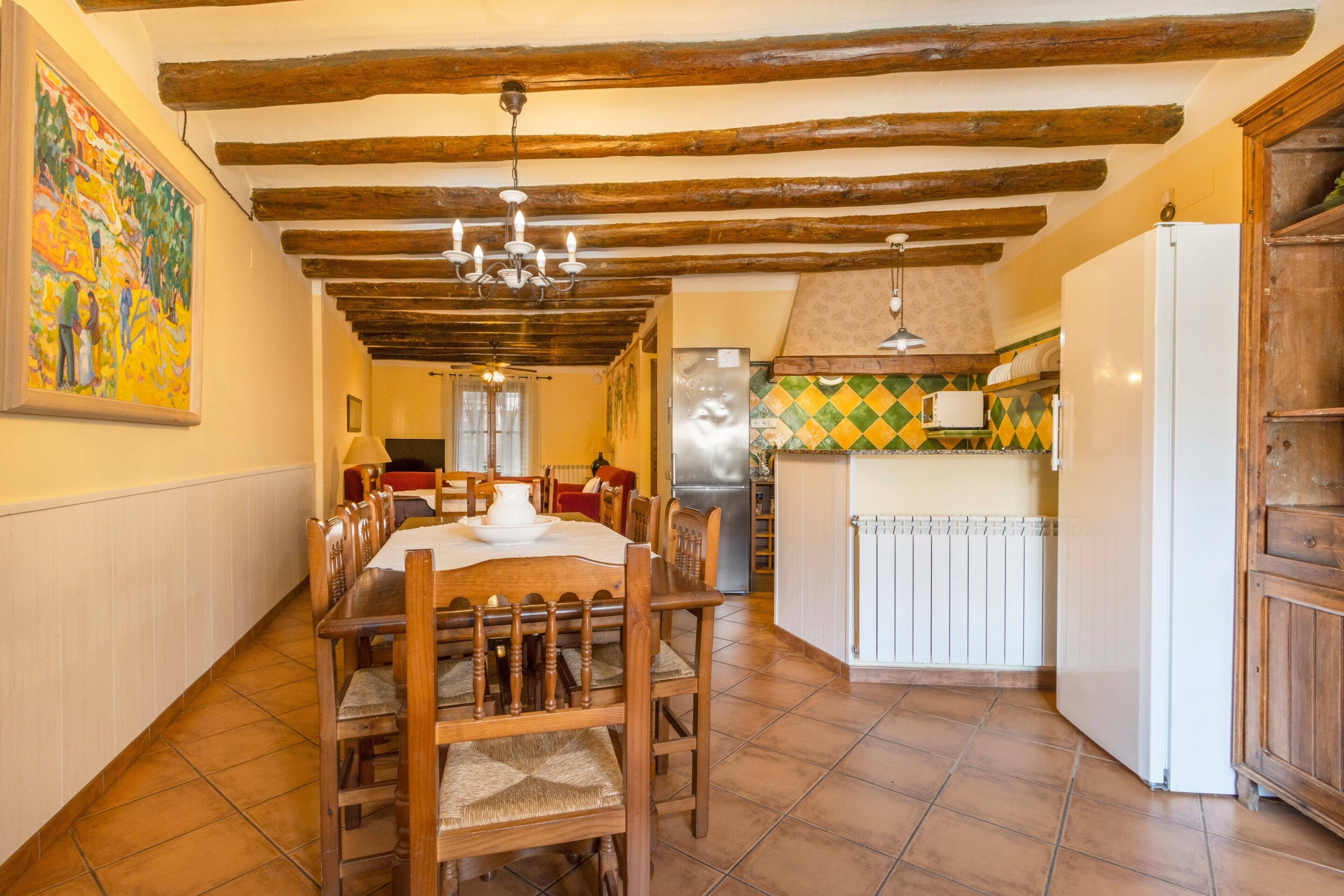 A dining area with wooden furniture, a chandelier, and a kitchen with a refrigerator and tiled backsplash.