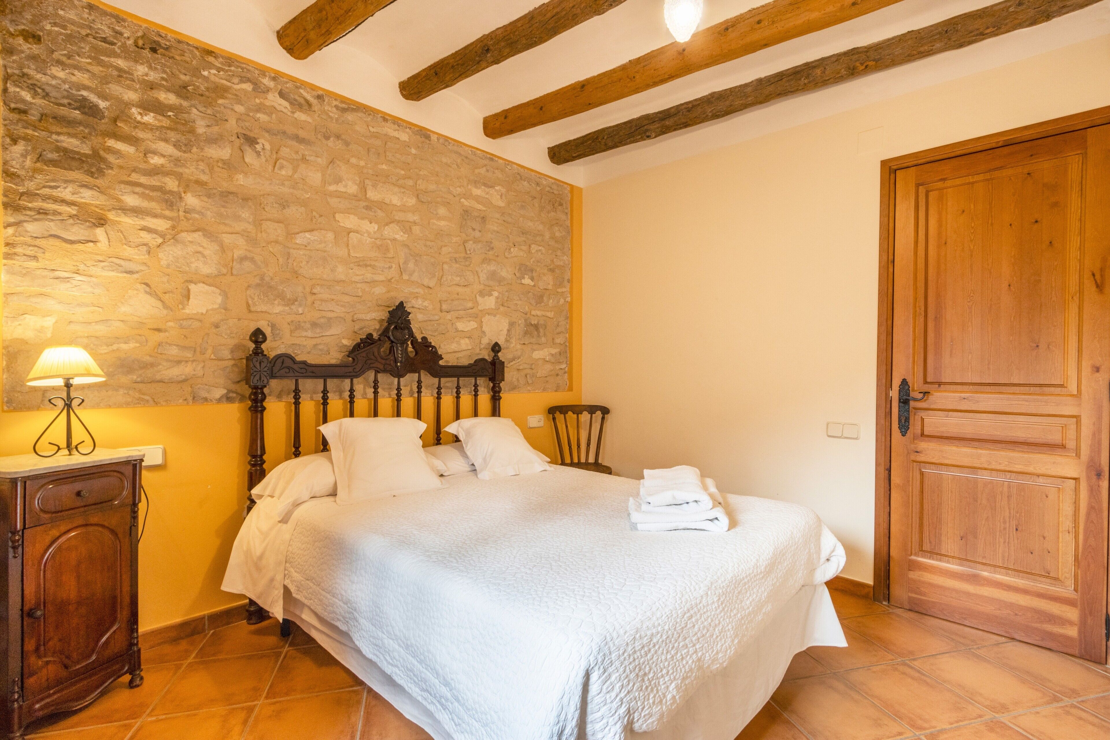 Un dormitorio con pared de piedra, vigas de madera en el techo, una cama con ropa blanca, una mesita de noche y una puerta de madera.