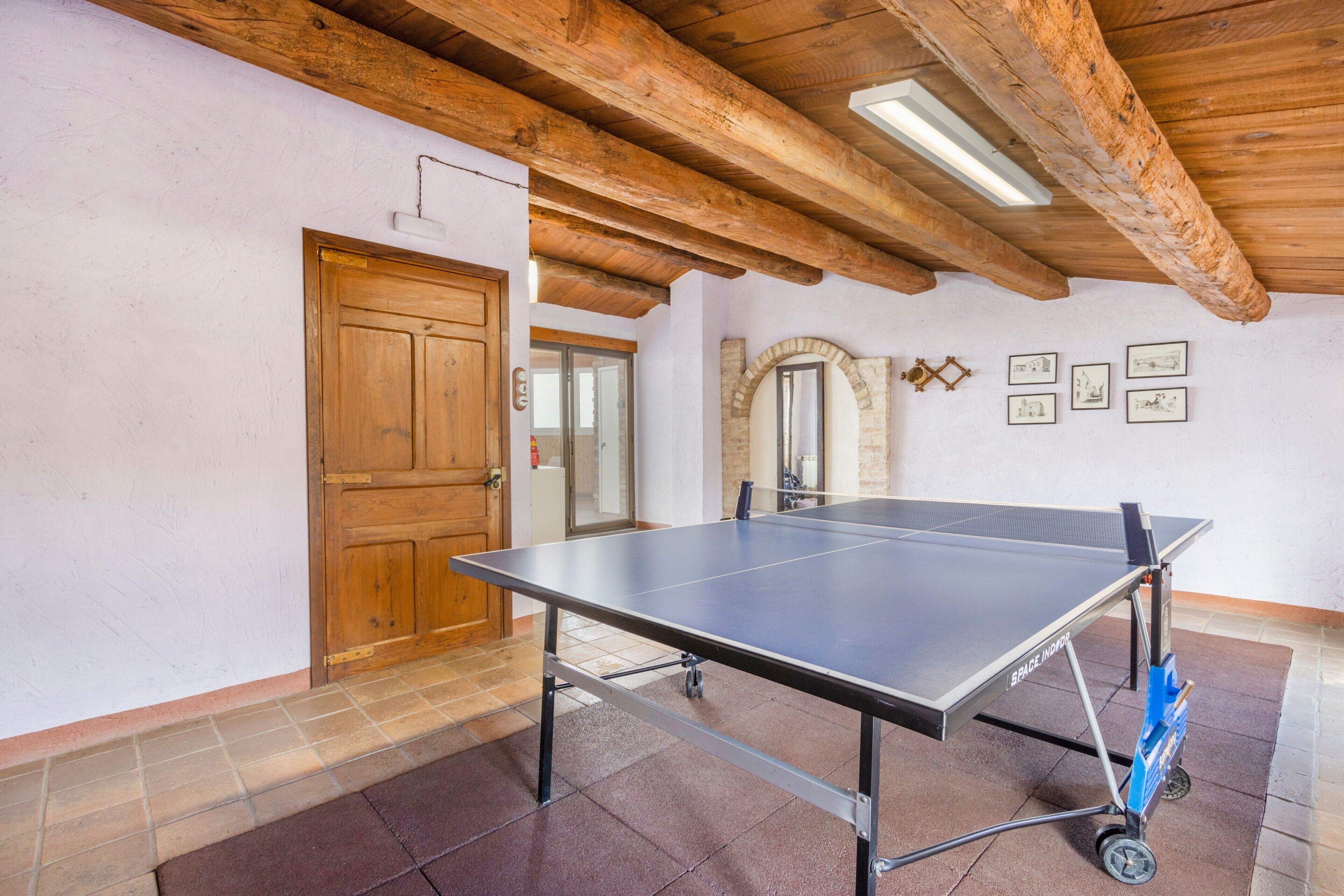 A ping pong table in a room with wooden beams, a door, and framed pictures on the wall.