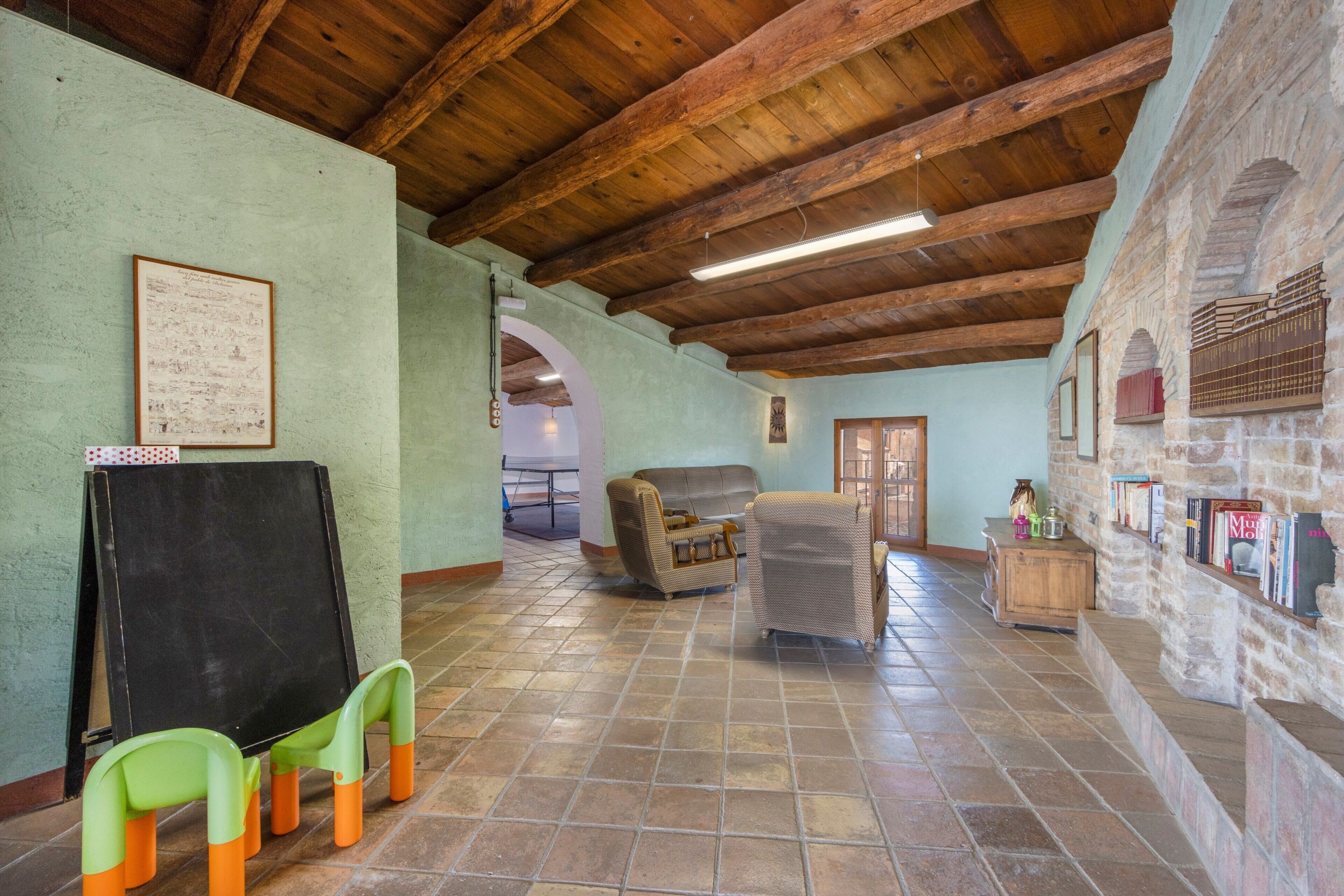 Una habitación amplia con un techo de madera alto, una pizarra, una silla verde, una silla de mimbre y una pared de piedra con estantes.
