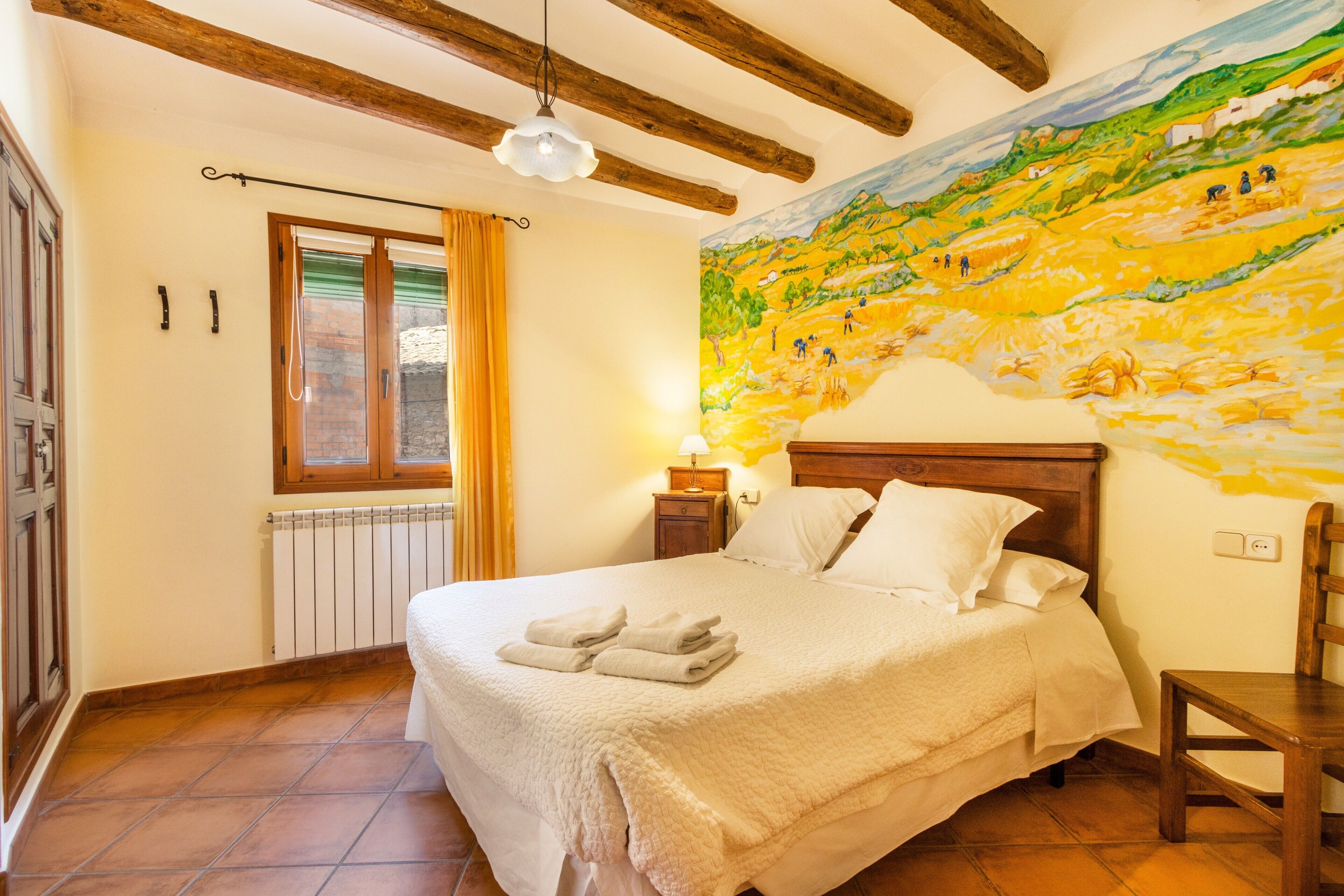 Un dormitori amb un llit gran, un panell de fusta al capçal, una petita taula de nit amb una làmpada, una finestra amb cortines i un mural a la paret.