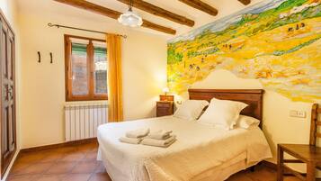 Un dormitori amb un llit gran, un panell de fusta al capçal, una petita taula de nit amb una làmpada, una finestra amb cortines i un mural a la paret.