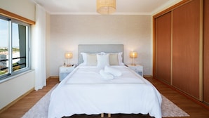 A neatly made bed with white linens, two bedside lamps, a large window, and a wooden wardrobe.