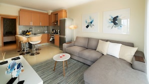A modern living room with a grey sofa, a small round table, and a kitchen area with wooden cabinets and a refrigerator.