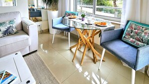 Een moderne woonkamer met een glazen eettafel, blauwe stoelen, een bank en een groot raam met gordijnen.