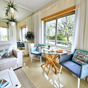 Un salon moderne avec une table à manger en verre, des chaises bleues, un canapé et une grande fenêtre avec des rideaux.