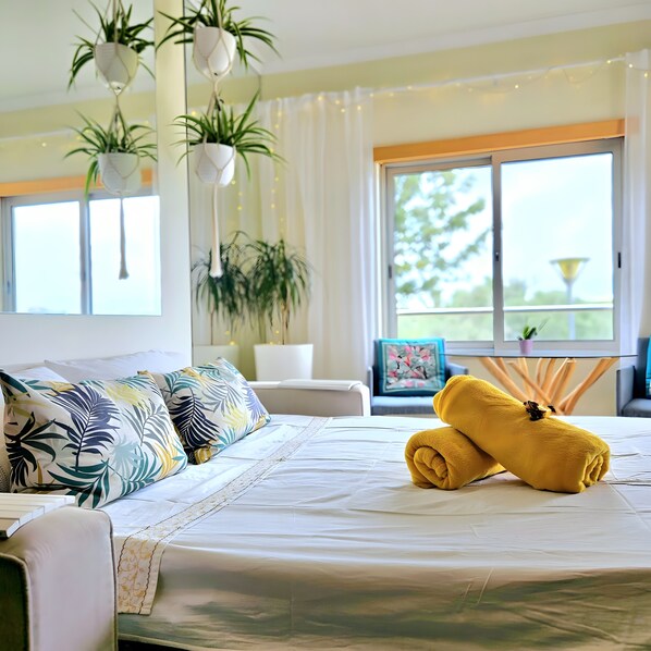 A hotel room with a bed, pillows, a yellow towel, hanging plants, a window, and a view of greenery.