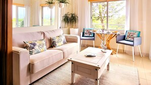 Een moderne woonkamer met een bank, salontafel en twee stoelen. Er zijn hangplanten, een raam met gordijnen en uitzicht op groen buiten.