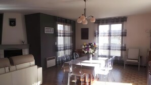 Ett modernt vardagsrum med en soffa, ett matbord, stolar, en öppen spis och stora fönster med gardiner.