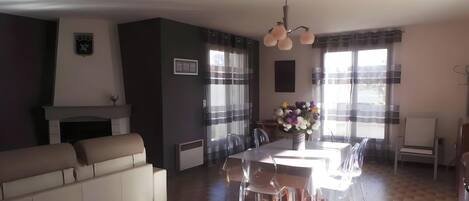Una sala de estar moderna con sofá, mesa de comedor, sillas, chimenea y ventanas grandes con cortinas.