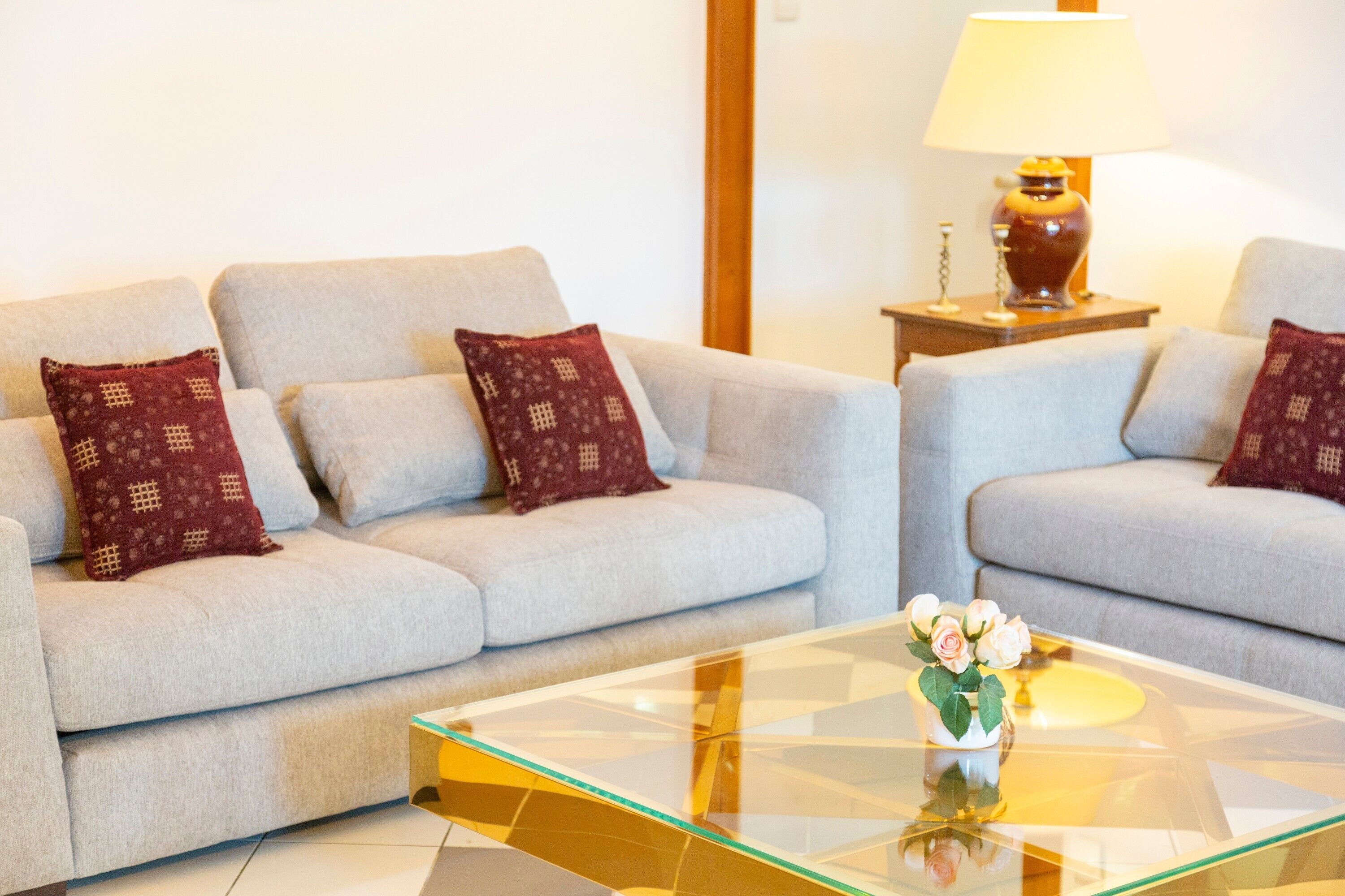 Un salon avec deux canapés gris clair, chacun agrémenté d’un coussin rouge à motifs, une table basse en verre avec un vase de fleurs et une lampe posée sur une table d’appoint en bois.