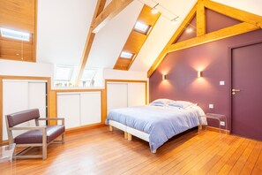 A bedroom with a sloped ceiling, wooden beams, a bed with a blue bedspread, a chair, and a door.