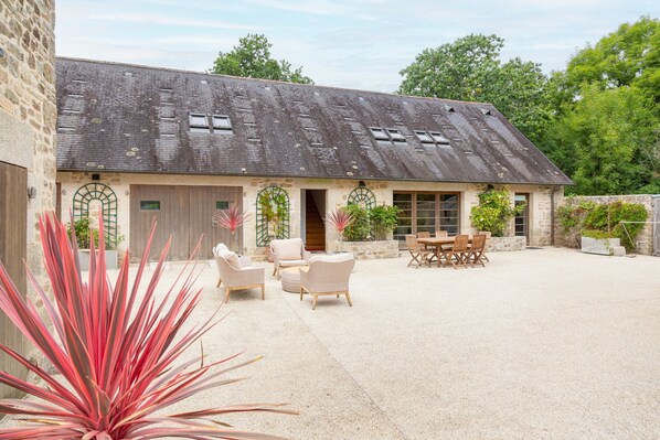 A stone building with a sloped roof, a covered patio with seating, and a large plant in the foreground.
