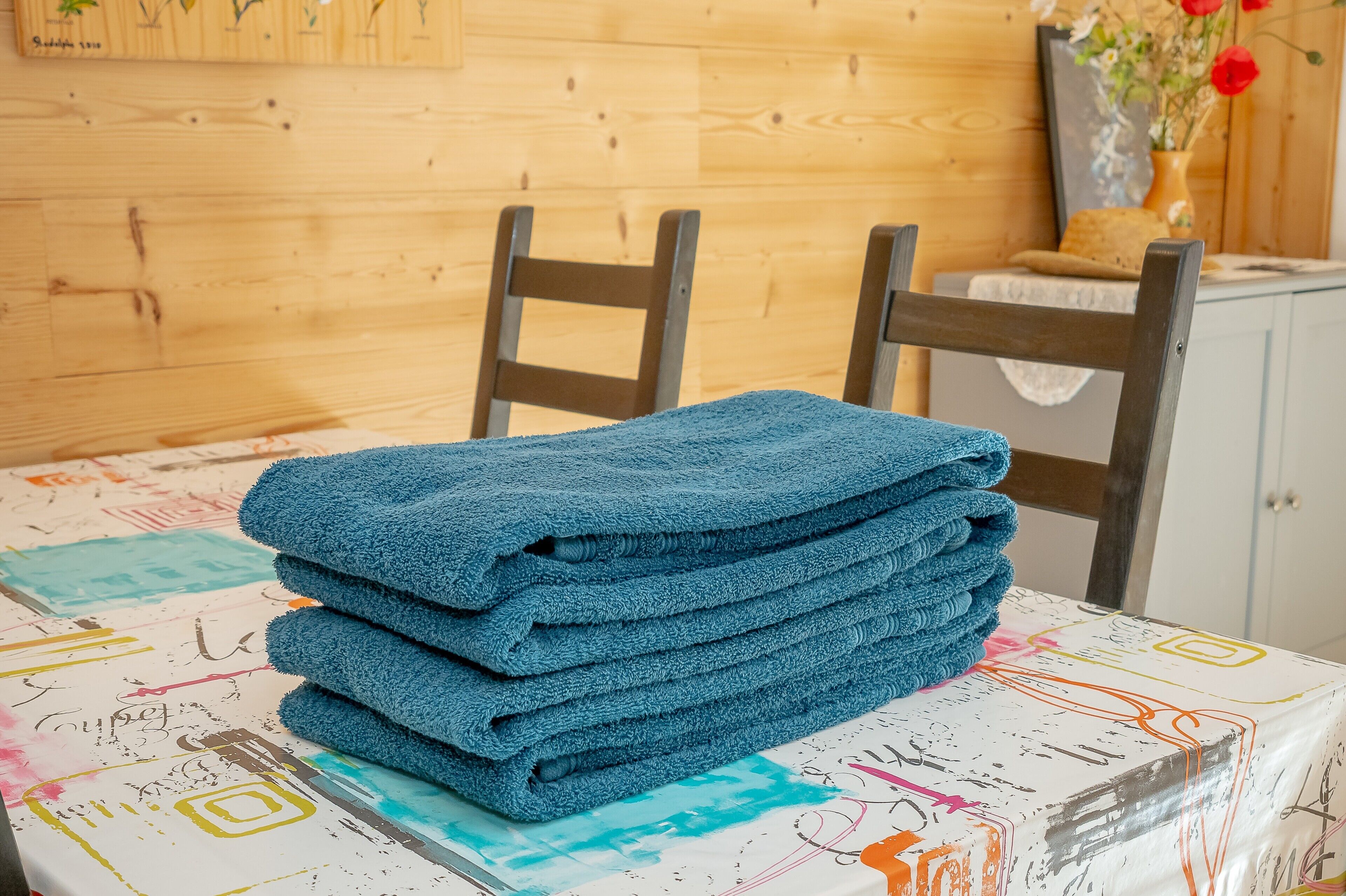 Ein Stapel blauer Handtücher auf einem Tisch mit farbenfroher Tischdecke, Holzsessel und einer Holzwand mit einem gerahmten Bild.
