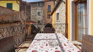 Un balcone con un tavolo apparecchiato per mangiare, una sedia in vimini e vista sugli edifici vicini.