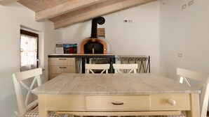 Zona pranzo con tavolo in legno, sedie bianche, forno a pizza e una finestra con tende.