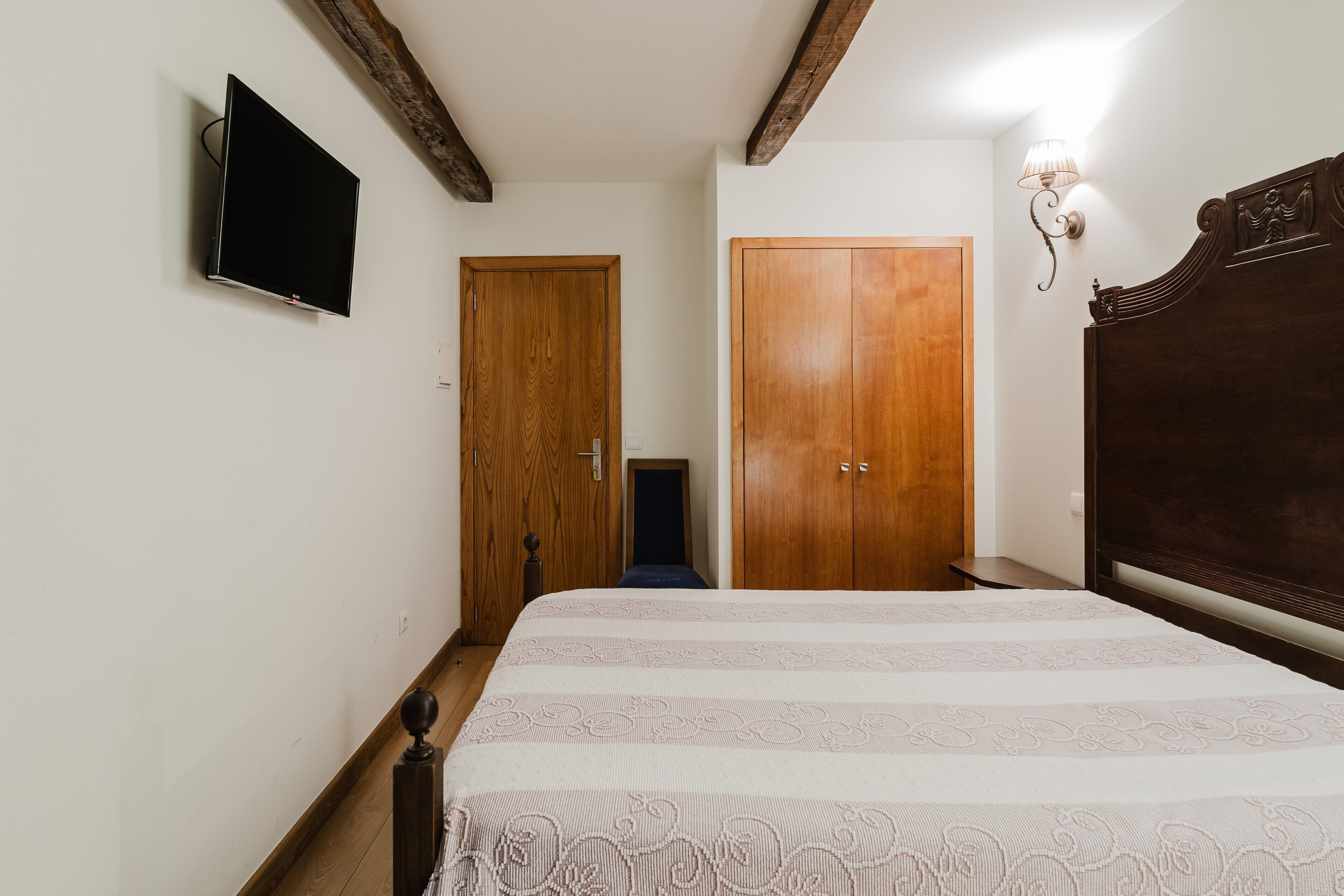 Un dormitorio con una cama, televisión montada en la pared, puertas de madera y una mesita de noche.