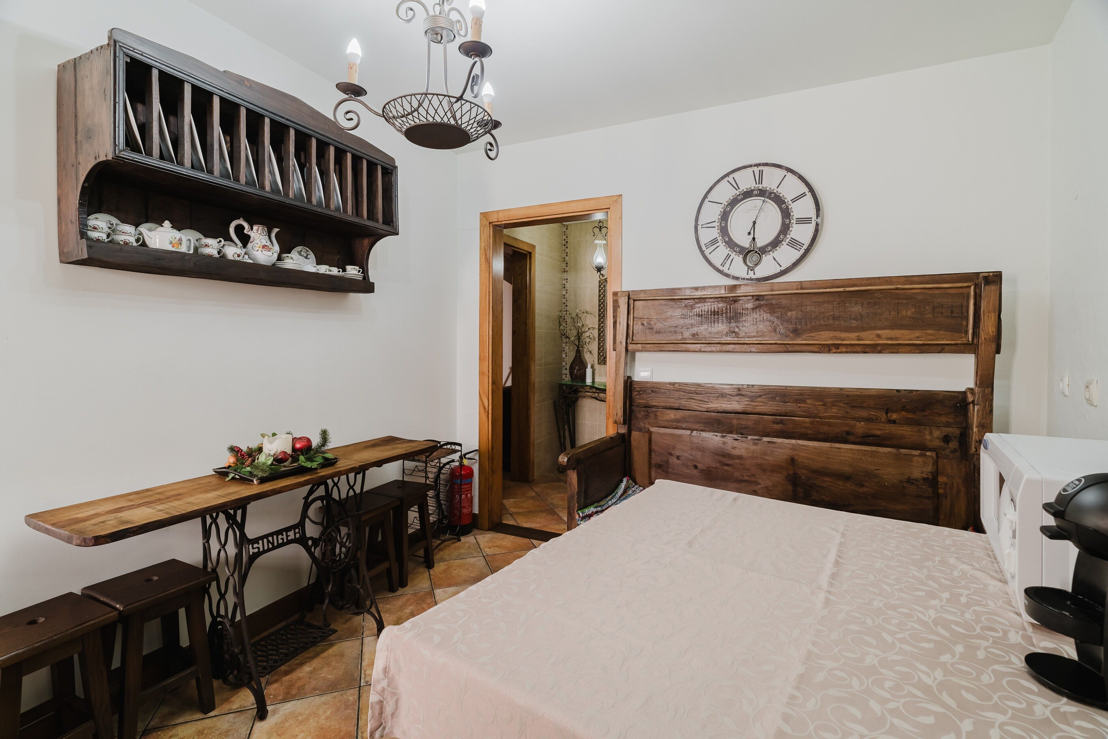 A bedroom with a wooden bed, a wooden desk, a shelf with decorative items, a wall clock, and a door leading to another room.